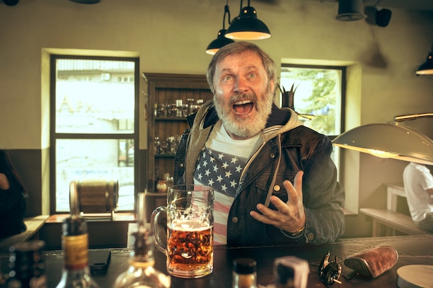 Senior homme barbu, boire de l'alcool dans un pub et regarder un programme sportif à la télévision.