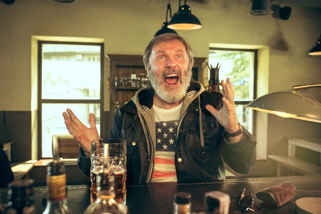 Senior homme barbu, boire de l'alcool dans un pub et regarder un programme sportif à la télévision.