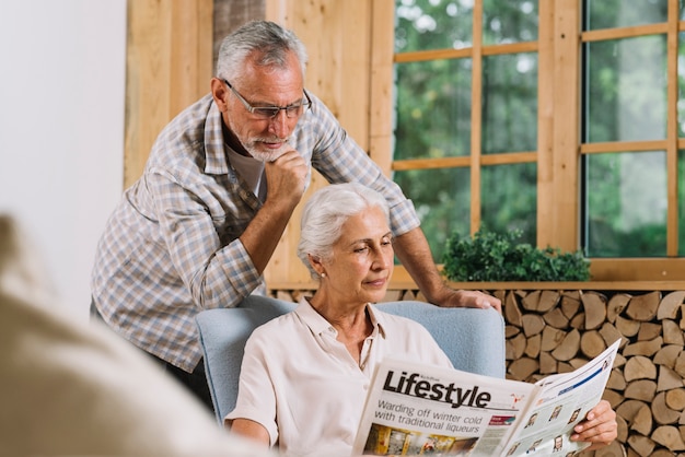 Senior homme debout derrière la femme assise sur une chaise lisant un journal