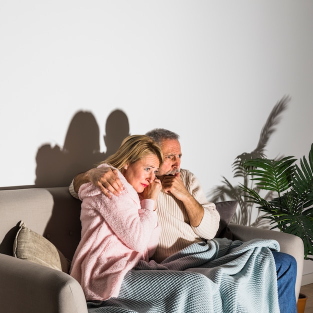 Senior homme effrayé étreignant avec femme et regarder la télévision sur un canapé