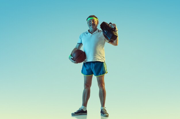 Senior homme posant de superbes vêtements de sport avec un magnétophone rétro sur un mur dégradé, néon. Modèle masculin caucasien en grande forme, sportif. Concept de sport, activité, mouvement, mode de vie sain.