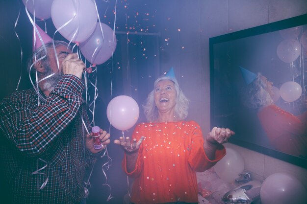 Senior homme souriant, soufflant une baguette de bulle et sa femme jetant des confettis à la fête d&#39;anniversaire