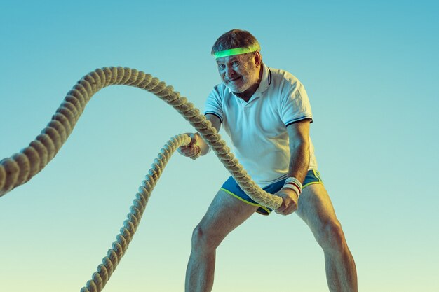 Senior man training avec des cordes sur un mur dégradé en néon. Le modèle masculin de race blanche en grande forme reste actif, sportif. Concept de sport, activité, mouvement, bien-être, mode de vie sain.