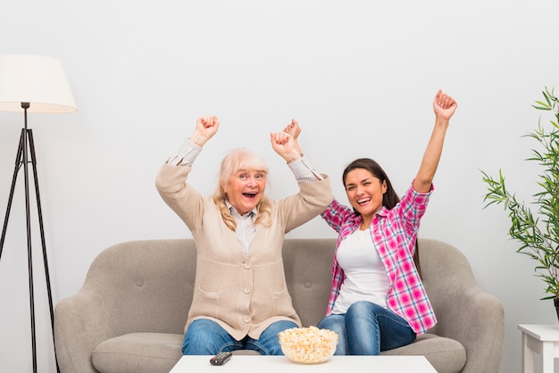 Senior mère excitée et sa fille assise sur un canapé en levant les bras en regardant la télévision