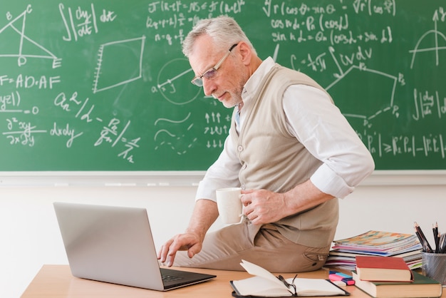 Senior professeur assis sur un bureau et en train de taper sur un ordinateur portable