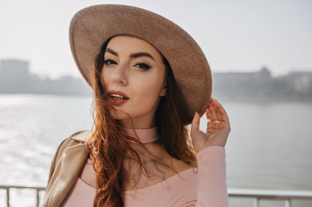 Sensuelle femme brune touchant son chapeau et regardant avec intérêt sur le mur de la rivière