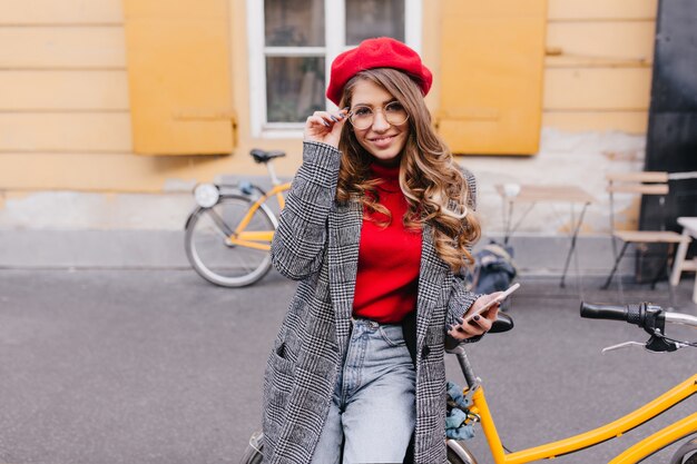 Sensuelle jeune femme blanche en long manteau vintage touchant ses grosses lunettes et souriant