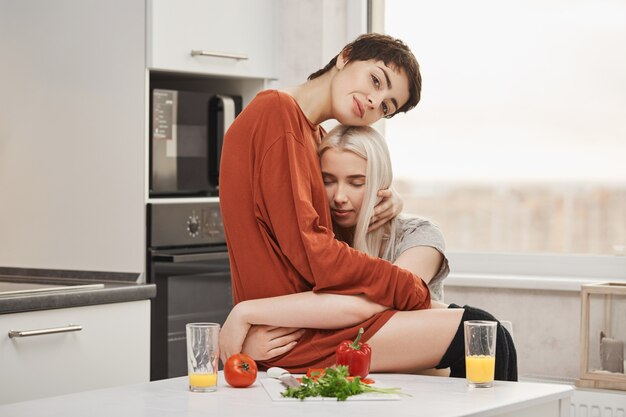 Sensuelle et tendre jeune couple de lesbiennes amoureux, câlins dans la cuisine tout en prenant le petit déjeuner. La femme est heureuse pour chaque jour qu'elle a passé avec sa petite amie, voulant l'étreindre et être avec elle pour toujours.