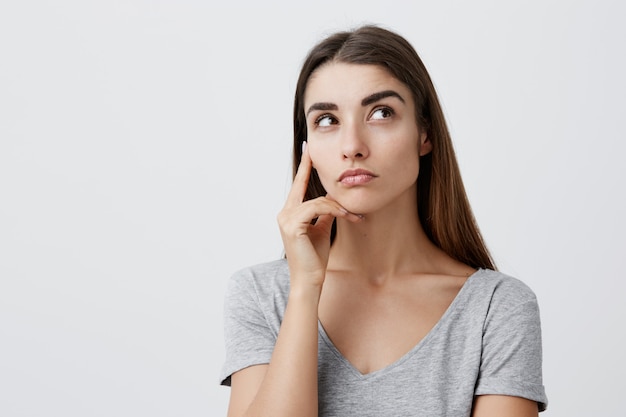 Sérieuse charmante belle brune étudiante caucasienne avec une longue coiffure en t-shirt gris élégant tenant la main sur la joue, regardant de côté avec une expression affectueuse et réfléchie,