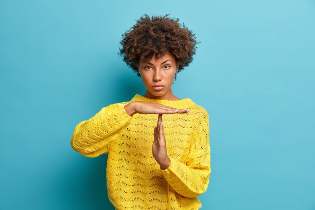 Sérieuse femme aux cheveux bouclés confiant fait un geste de dépassement de temps montre limite demande d'arrêter habillé en pull jaune isolé sur mur bleu