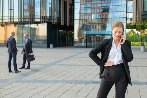 Sérieuse jeune femme d'affaires en costume de bureau parlant au téléphone portable à l'extérieur. Les gens d'affaires et la façade en verre du bâtiment de la ville en arrière-plan. Copiez l'espace. Concept de communication d'entreprise