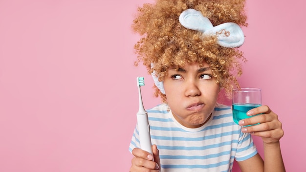 Photo gratuite sérieuse jeune femme aux cheveux bouclés concentrée de côté utilise un rince-bouche et une brosse à dents électrique nettoie les dents après le réveil porte un t-shirt rayé isolé sur fond rose espace vide pour votre texte