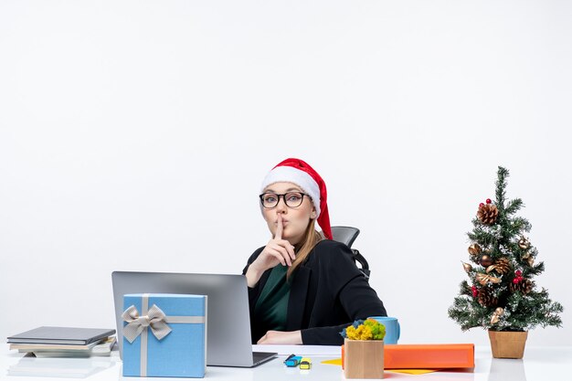 Sérieuse jeune femme avec chapeau de père Noël et lunettes assis à une table avec un arbre de Noël et un cadeau dessus faisant le geste de silence sur fond blanc