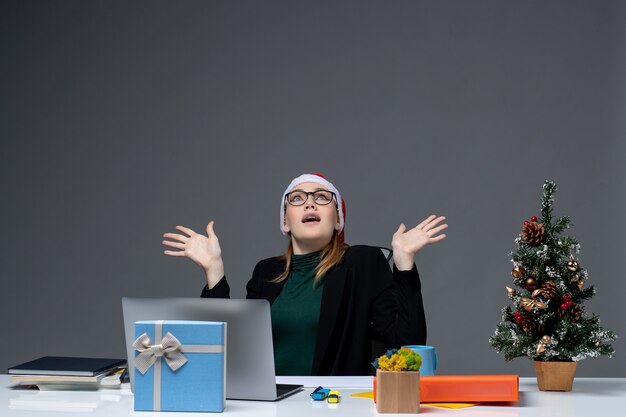 Sérieuse jeune femme avec chapeau de père Noël et lunettes assis à une table avec un arbre de Noël et un cadeau dessus sur fond sombre