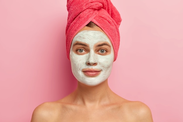 Sérieuse jeune femme avec masque facial à l'argile, porte une serviette enveloppée, nourrit la peau avec des vitamines