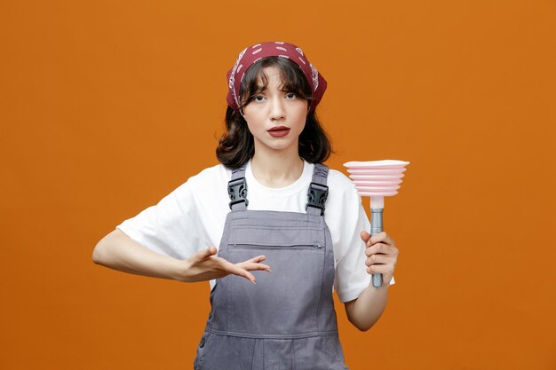 Sérieuse jeune femme nettoyante portant l'uniforme et le bandana montrant le piston pointant dessus avec les mains regardant la caméra isolée sur fond orange