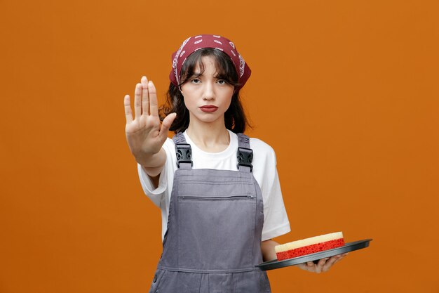 Sérieuse jeune femme nettoyante portant un uniforme et un bandana tenant un plateau avec une éponge dedans regardant la caméra montrant un geste d'arrêt isolé sur fond orange