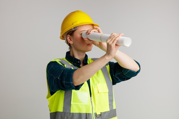 Sérieuse jeune femme travailleur de la construction portant un casque de sécurité et un gilet de sécurité tenant du papier à l'aide comme télescope à côté