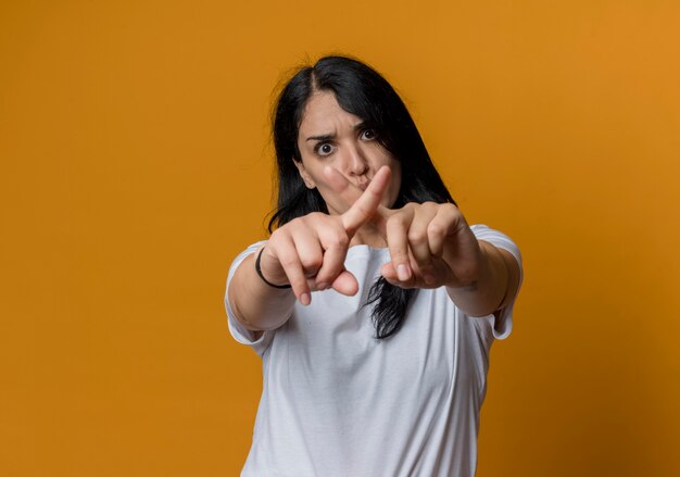 Sérieuse jeune fille caucasienne brune croise les doigts ne faisant aucun signe isolé sur un mur orange