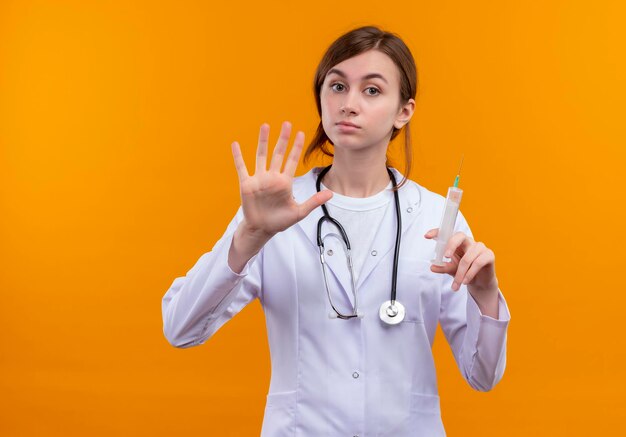 Sérieusement à la jeune femme médecin portant une robe médicale et un stéthoscope tenant une seringue et montrant cinq sur l'espace orange isolé avec copie espace