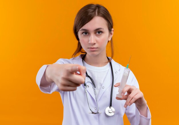 Sérieusement à la jeune femme médecin portant robe médicale et stéthoscope tenant la seringue et pointant sur l'espace orange isolé