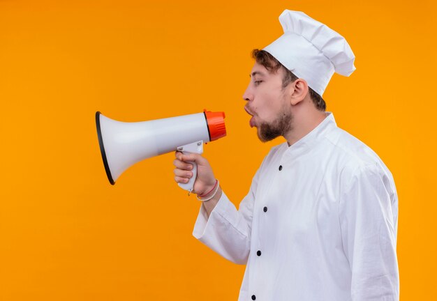 Un sérieux beau jeune chef barbu homme en uniforme blanc parlant à travers un mégaphone sur un mur orange