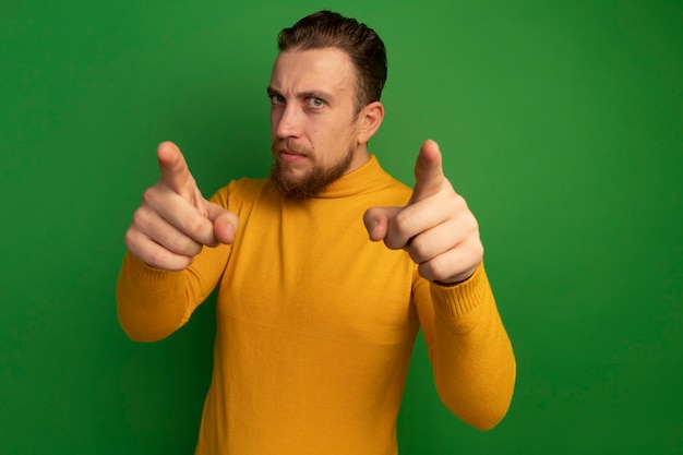 Photo gratuite sérieux bel homme blond pointe à l'avant avec deux mains isolé sur mur vert