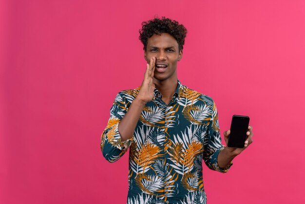 Sérieux et confiant beau homme à la peau sombre aux cheveux bouclés appelant quelqu'un à distance tenant un téléphone mobile sur fond rose