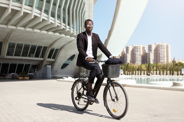 Sérieux et confiant jeune homme afro-américain employé de bureau portant des lunettes de soleil en miroir et cyclisme costume noir formel