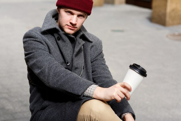 Sérieux garçon au chapeau rouge est assis avec une tasse de café dans la rue