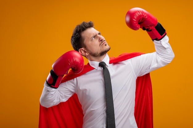 Sérieux homme d'affaires de super héros confiant en cape rouge et en gants de boxe levant les mains montrant la force et le courage debout sur fond orange