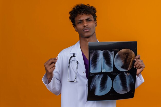 Un sérieux jeune beau médecin de sexe masculin à la peau sombre avec des cheveux bouclés portant blouse blanche avec stéthoscope montrant le rapport de rayons x