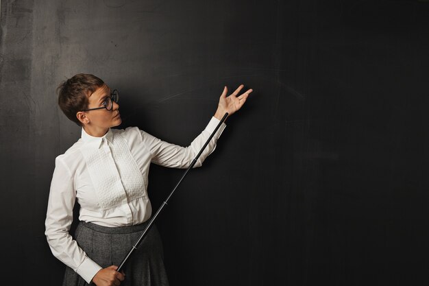 Sérieux à la jeune enseignante blanche en blanc bouton blouse et jupe en tweed se dresse à un tableau noir tenant un pointeur