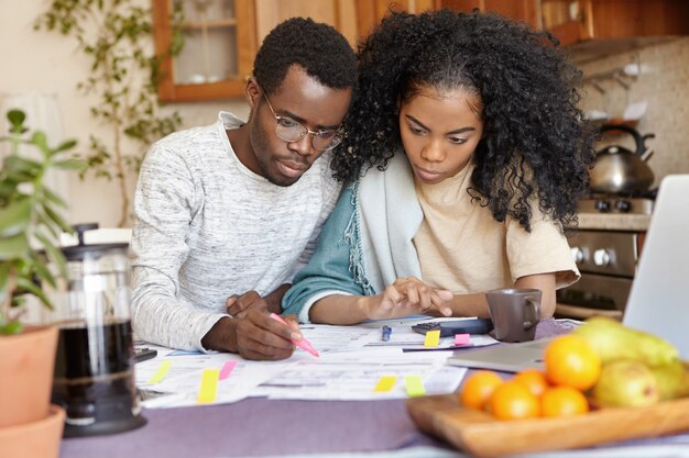 Sérieux jeune homme africain dans des verres tenant un stylo-feutre, calculant les dépenses domestiques tout en gérant le budget familial avec sa belle femme, en planifiant un gros achat et en essayant d'économiser de l'argent