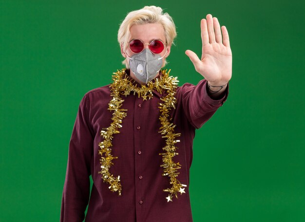 Photo gratuite sérieux jeune homme blond portant des lunettes et un masque de protection avec guirlande de guirlandes autour du cou faisant le geste d'arrêt isolé sur mur vert