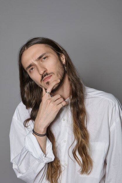 Sérieux jeune homme hipster aux cheveux longs réfléchi portant une chemise blanche élégante, touchant la barbe, ayant un regard pensif profondément dans ses pensées, ses yeux pleins de doute et d'incertitude. Expression faciale humaine