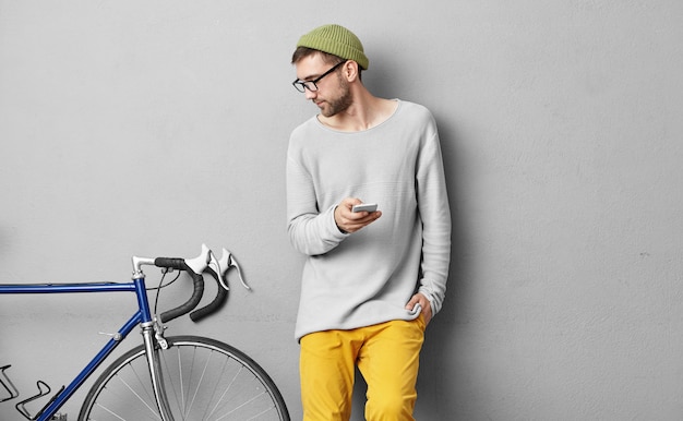 Sérieux jeune homme mal rasé dans des vêtements à la mode posant sur un mur de béton et regardant son vélo à pignon fixe, étudiant ses caractéristiques tout en publiant une annonce via un site Web classé, en le mettant en vente