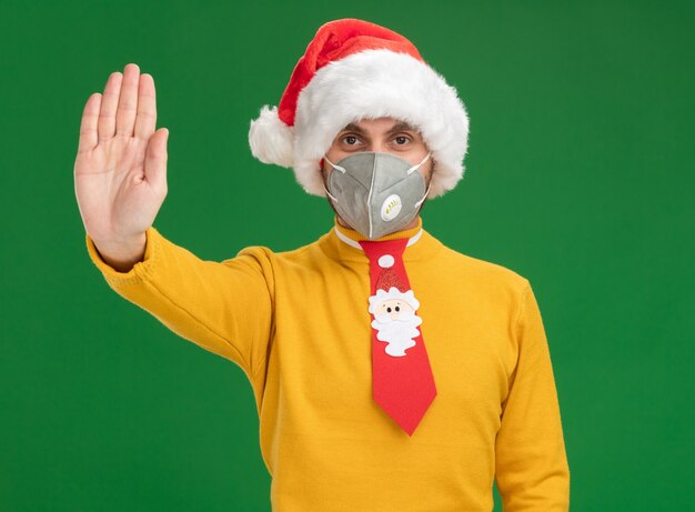 Sérieux jeune homme de race blanche portant chapeau de Noël et cravate avec masque de protection regardant la caméra faisant le geste d'arrêt isolé sur fond vert