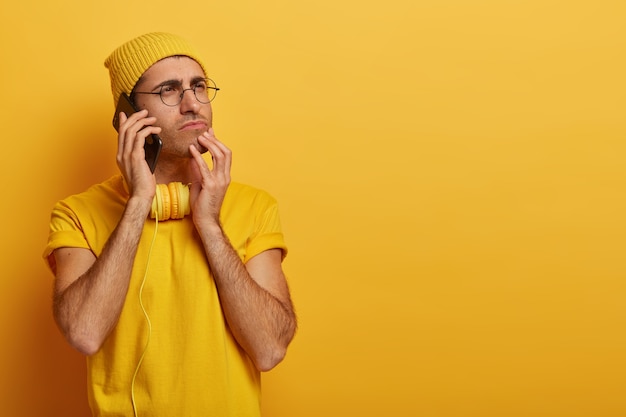 Sérieux jeune homme de race blanche songeur se détourne, pense à quelque chose de profondément, porte des lunettes transparentes, un t-shirt jaune décontracté et un couvre-chef