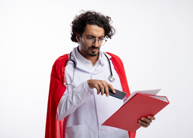 Sérieux jeune homme de super-héros caucasien à lunettes optiques portant l'uniforme de médecin avec manteau rouge et avec stéthoscope autour du cou tient et pointe au dossier avec téléphone avec espace de copie