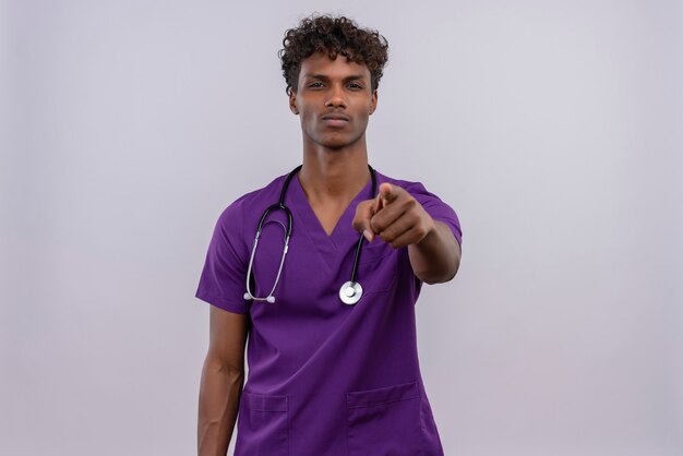 Sérieux jeune médecin de sexe masculin à la peau sombre avec des cheveux bouclés portant l'uniforme violet avec stéthoscope pointant avec l'index à la caméra sur un fond blanc