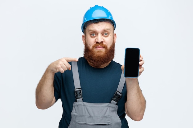 Sérieux jeune ouvrier du bâtiment masculin portant un casque de sécurité et un uniforme regardant la caméra montrant un téléphone portable à la caméra pointant dessus isolé sur fond blanc