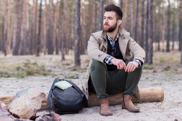 Photo gratuite sérieux jeune randonneur avec son sac à dos, assis sur un journal à la plage