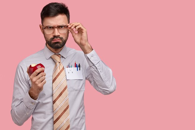 Sérieux patron masculin sombre dans des vêtements formels garde la main sur des lunettes, mord une délicieuse pomme rouge