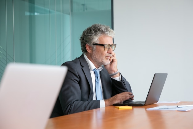 Sérieux professionnel mature en costume et lunettes parler au téléphone portable, travaillant à l'ordinateur portable au bureau, regardant l'affichage