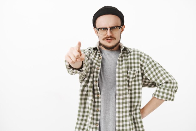 Sérieux à la stricte, irrité jeune professeur de musique de sexe masculin en bonnet noir et lunettes avec moustache debout dans une posture furieuse pointant vers l'avant