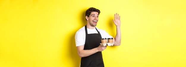 Serveur amical au café agitant la main au client tenant du café à emporter debout contre le jaune