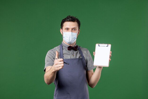 serveur de banquet en uniforme avec masque médical et tenant un carnet de chèques faisant un geste correct sur fond vert