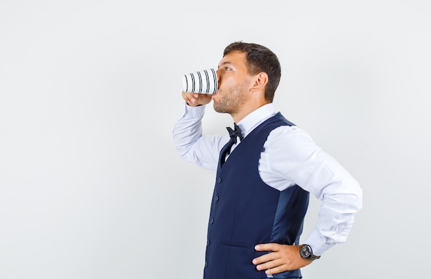 Serveur buvant une tasse de thé en chemise blanche, gilet bleu foncé.