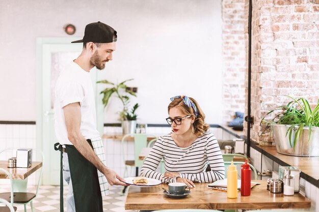 Serveur en casquette noire donnant un hamburger à une jolie dame à lunettes assise à la table du café